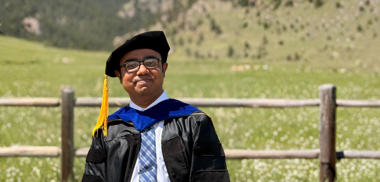 Dipto is wearing a graduation gown outside with a green background of a blurred out field behind him.