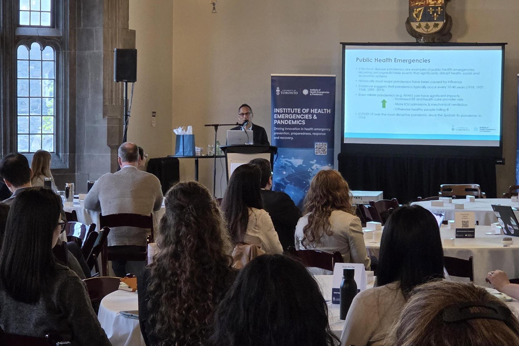 Howard Njoo presenting at a podium during an Institute of Health Emergencies & Pandemics event, addressing an audience seated at round tables. A projected slide titled “Public Health Emergencies” and an institute banner are visible behind the speaker, with large windows and a historic hall setting in the background.